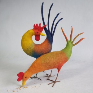 A wet felted cockerel with yellow and blue body stands behind a felted hen in shades of orange, red and green.